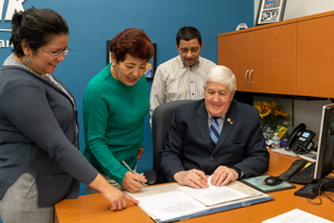 man seated at a desk reviewing a document with two women and a man standing near him looking down at the document