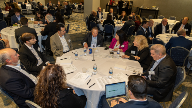 A conference room full of tables set with white tablecloths with a mix of men and women wearing professional attire sitting next to each other as they chat