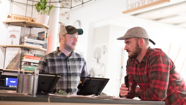 man standing in front of laptop screen while speaking to another man who is leaning on a counter