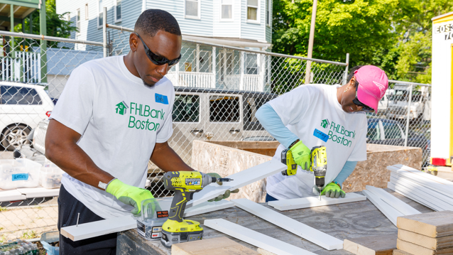 A man wearing protective gloves holding a strip of white painted wood standing next to a woman wearing protective gloves who is using a power drill on a strip of white painted wood