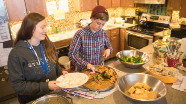 woman standing with an empty plate next to woman who is stirring food in a pan