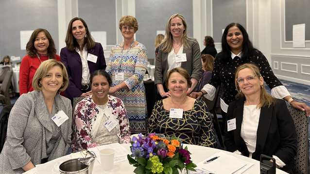 A group of women seated at a table next to each other smiling with another group of smiling women standing behind them.