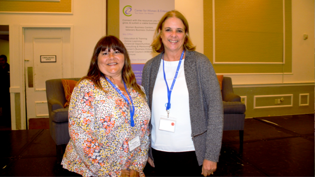 two female FHLBank Boston employees standing side by side smiling in front of a banner at a women's conference