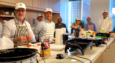Six FHLBank Boston senior leaders wearing white hats with FHLBank Boston logos standing in a kitchen in front of a long counter with electric skillets