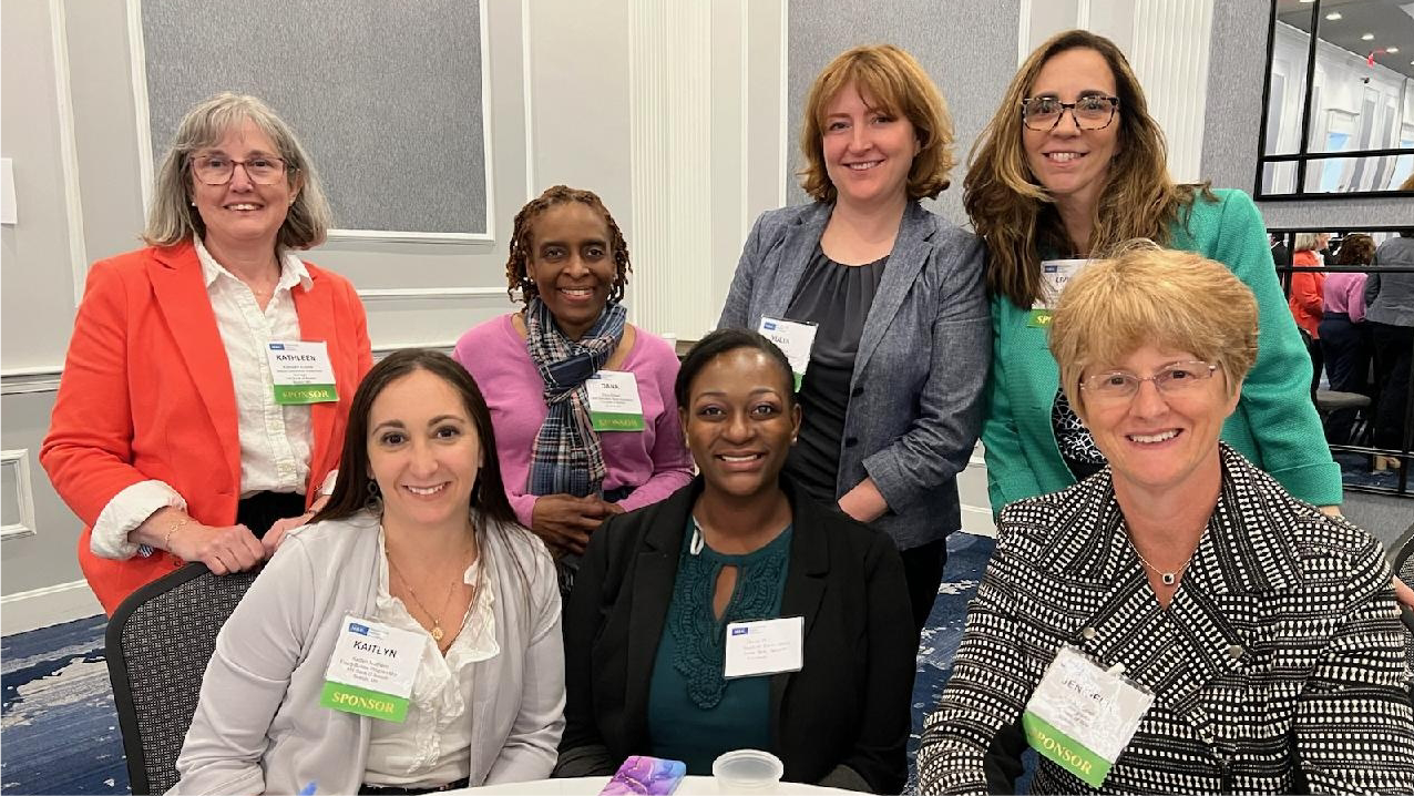 Group of seven female FHLBank Boston employees with four standing next to each other behind three that are seated at a round table.