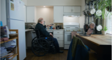 Man in wheelchair by the sink area of a kitchen with his hand placed firmly on the top of on insulated coffee mug. A laptop sits on a table to his right with pots and pans hanging on a rack near it.