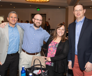A male FHLBank Boston employee standing between a male and female employee with his arms around their shoulders along with a fourth male employee standing next to the three.