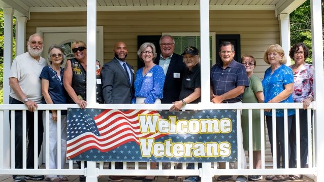 a mix of five men and six women standing next to each other on a porch with a welcome veterans banner draped over the porch railing