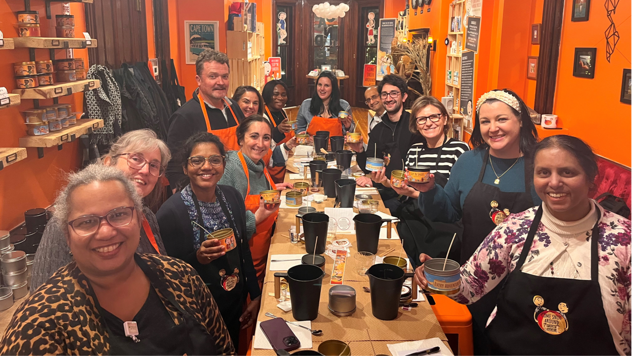 Mix of female and male FHLBank Boston employees standing around a long rectangular table with candle making supplies, with some employees holding candle canisters.