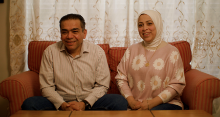 A smiling married couple sits on a sofa in the new home they purchased with the help of funding from an FHLBank Boston homebuyer assistance program.