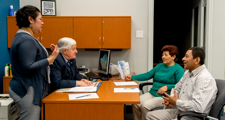 female standing near desk with man seated at desk and male and female couple seated in front of desk looking at seated male