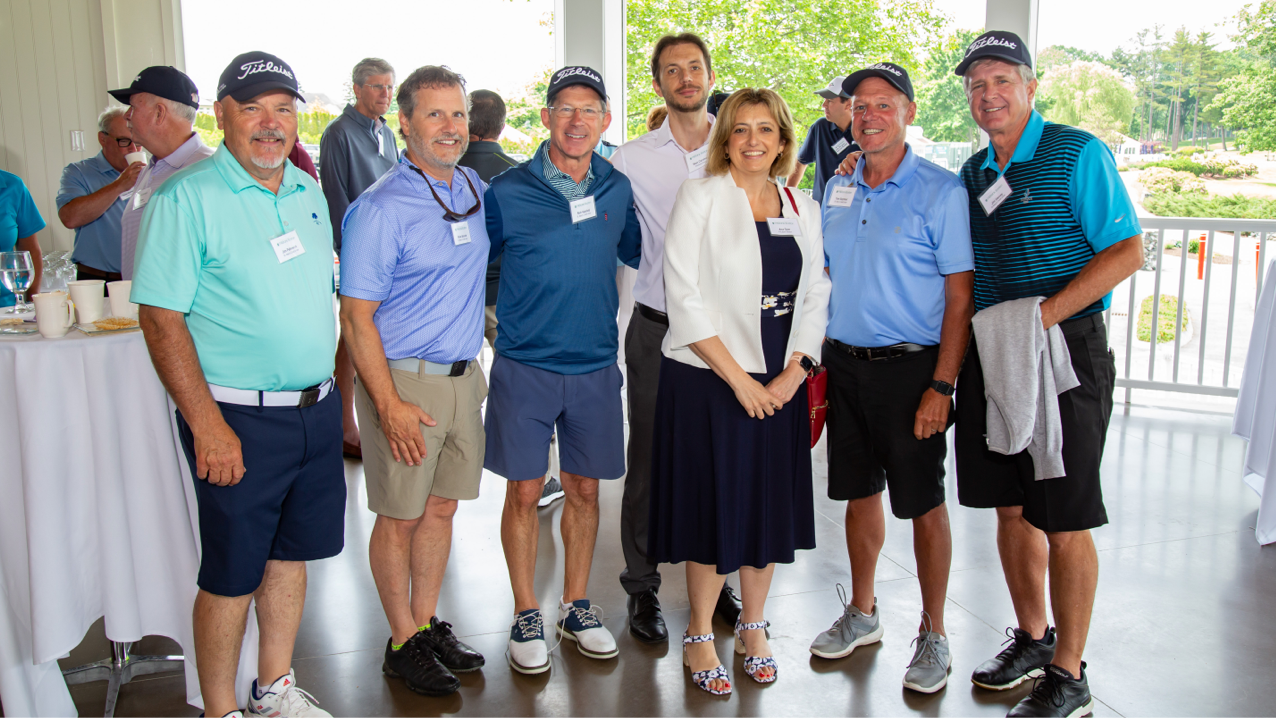 Six men wearing golf apparel, along with one woman, standing next to each other in a bright clubhouse room.