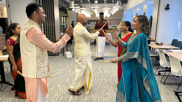Three men and three women wearing traditional Indian clothing standing in a café with two of the men and two of the women dancing as they hold gold wands
