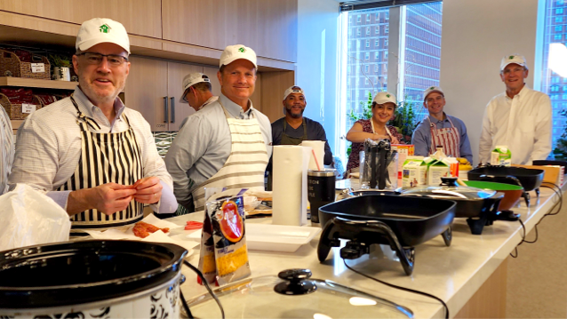 Six FHLBank Boston senior leaders wearing white hats with FHLBank Boston logos standing in a kitchen in front of a long counter with electric skillets