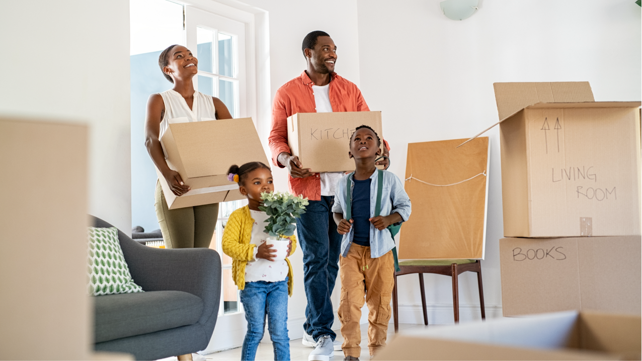 Adult man and woman holding moving boxes with a girl holding a plant and a boy standing in front of them as they walk into a home.