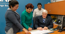 man seated at a desk reviewing a document with two women and a man standing near him looking down at the document