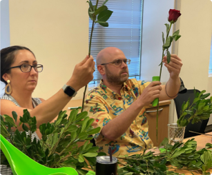 A female and male employee seated next to each other at a table with greens looking at red stem roses that they’re holding.