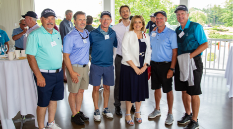 Six men wearing golf apparel, along with one woman, standing next to each other in a bright clubhouse room.