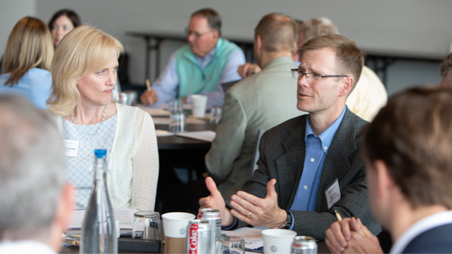 Woman looking at man seated next to her at a table who is talking to a group at the table.