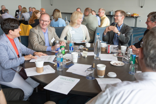 mix of women and men sitting at a round table with papers interacting and talking with each other