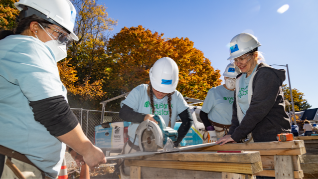 Four FHLBank Boston female employees wearing hardhats and protective googles standing outdoors watching as one of them uses an electric saw to cut a piece of wood