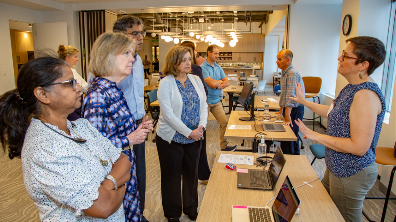 Three female FHLBank Boston employees and a male employee standing next to each other listening attentively in front of a table with 2 laptops as a woman behind the table explains the technology