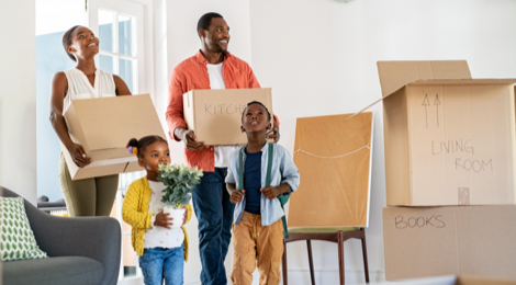 Adult man and woman holding moving boxes with a girl holding a plant and a boy standing in front of them as they walk into a home.