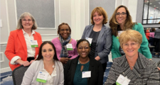 Group of seven female FHLBank Boston employees with four standing next to each other behind three that are seated at a round table.