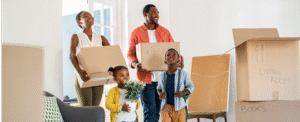 Adult man and woman holding moving boxes with a girl holding a plant and a boy standing in front of them as they walk into a home.