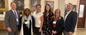 Four women standing between two men in a hallway of the Massachusetts State House with framed portraits of state leaders on the wall behind them.