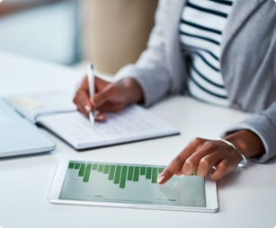 hands of a woman seated at a desk with a laptop as she writes in an open notebook and points to a tablet at her side