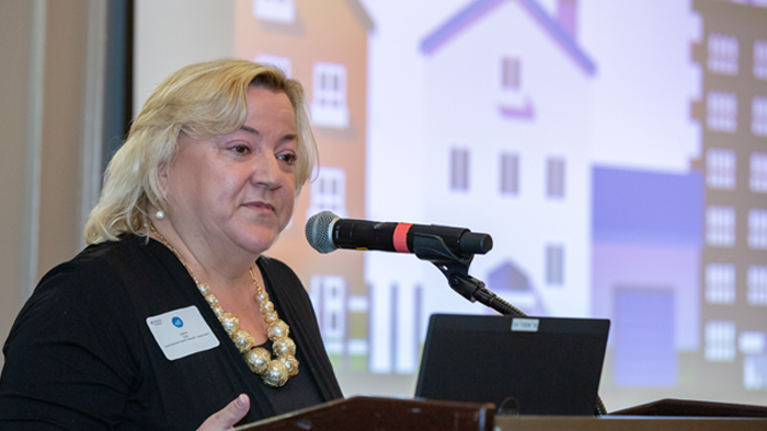 woman speaking at a podium with a microphone with a screen displayed behind her