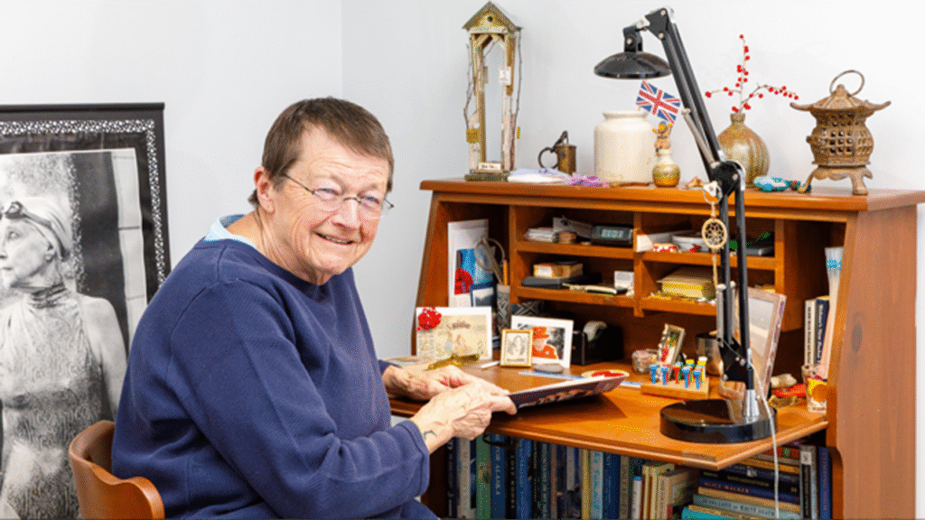 elderly woman wearing glasses seated at desk filled with knick-knacks as she holds a paper