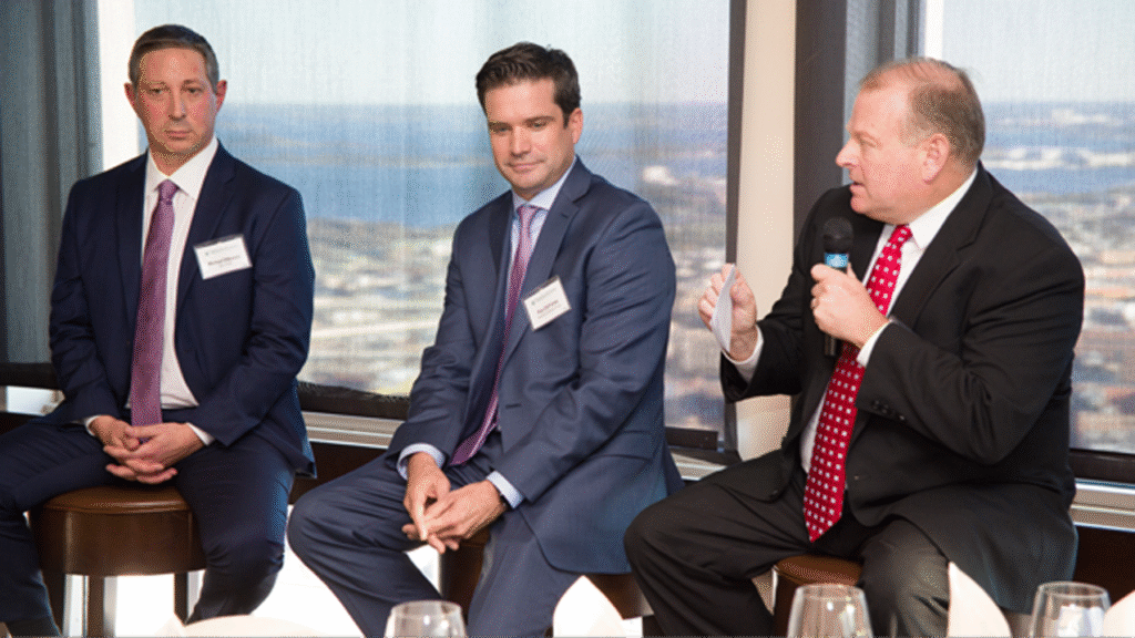 three male members seated as part of a panel speaking at an insurance company meeting with man at the right holding microphone
