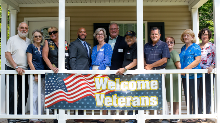 a mix of five men and six women standing next to each other on a porch with a welcome veterans banner draped over the porch railing
