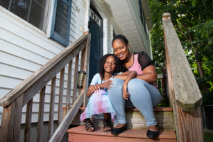 smiling mother and her young daughter sitting close to each other on the steps leading to the porch and front door of their home