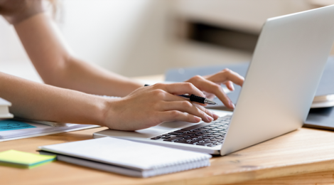 female hands typing at laptop while one hand holds a pen with a notebook lying on the desk next to the laptop