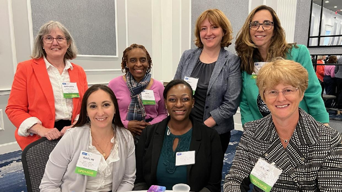Group of seven female FHLBank Boston employees with four standing next to each other behind three that are seated at a round table.

