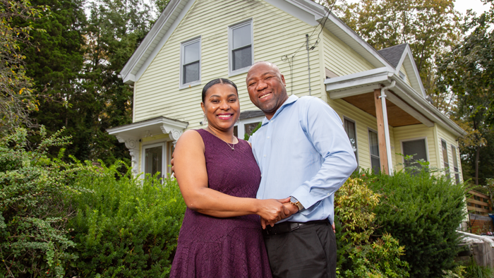Woman and man standing next to each other outdoors in front of their home while holding hands and smiling.