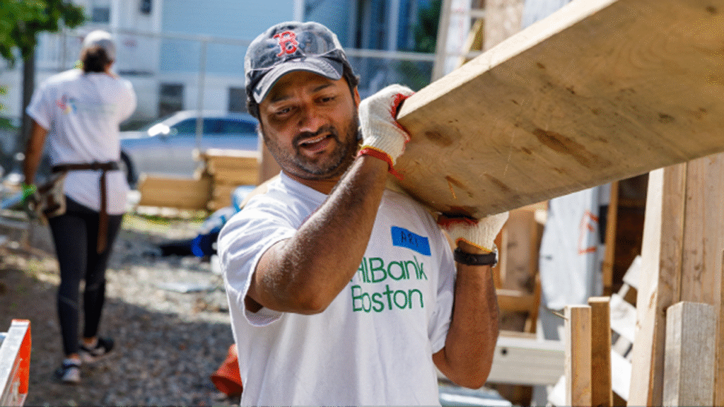 man wearing FHLBank Boston t-shirt holding a large piece of plywood by his shoulders as he stands at a construction site