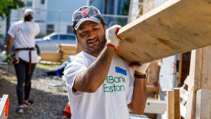 man wearing FHLBank Bosotn t-shirt holding a large piece of plywood at his shoulders as he stands at construction site
