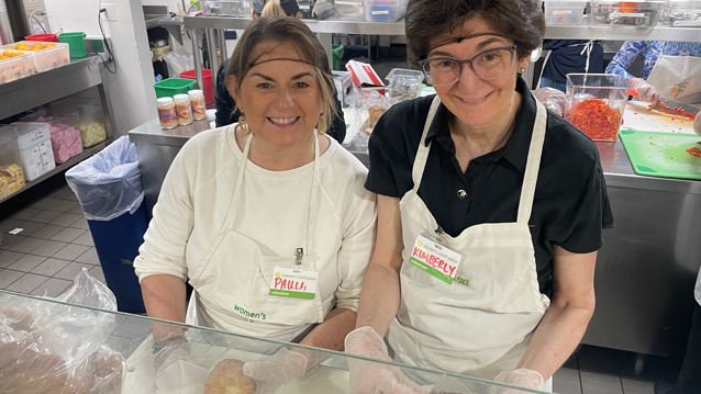 Two women wearing aprons, hairnets, and plastic gloves wrapping food at a commercial kitchen counter.