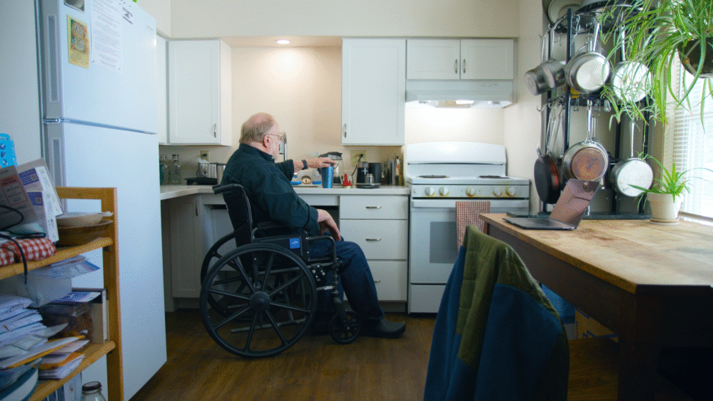 Man in wheelchair by the sink area of a kitchen with his hand placed firmly on the top of on insulated coffee mug. A laptop sits on a table to his right with pots and pans hanging on a rack near it.