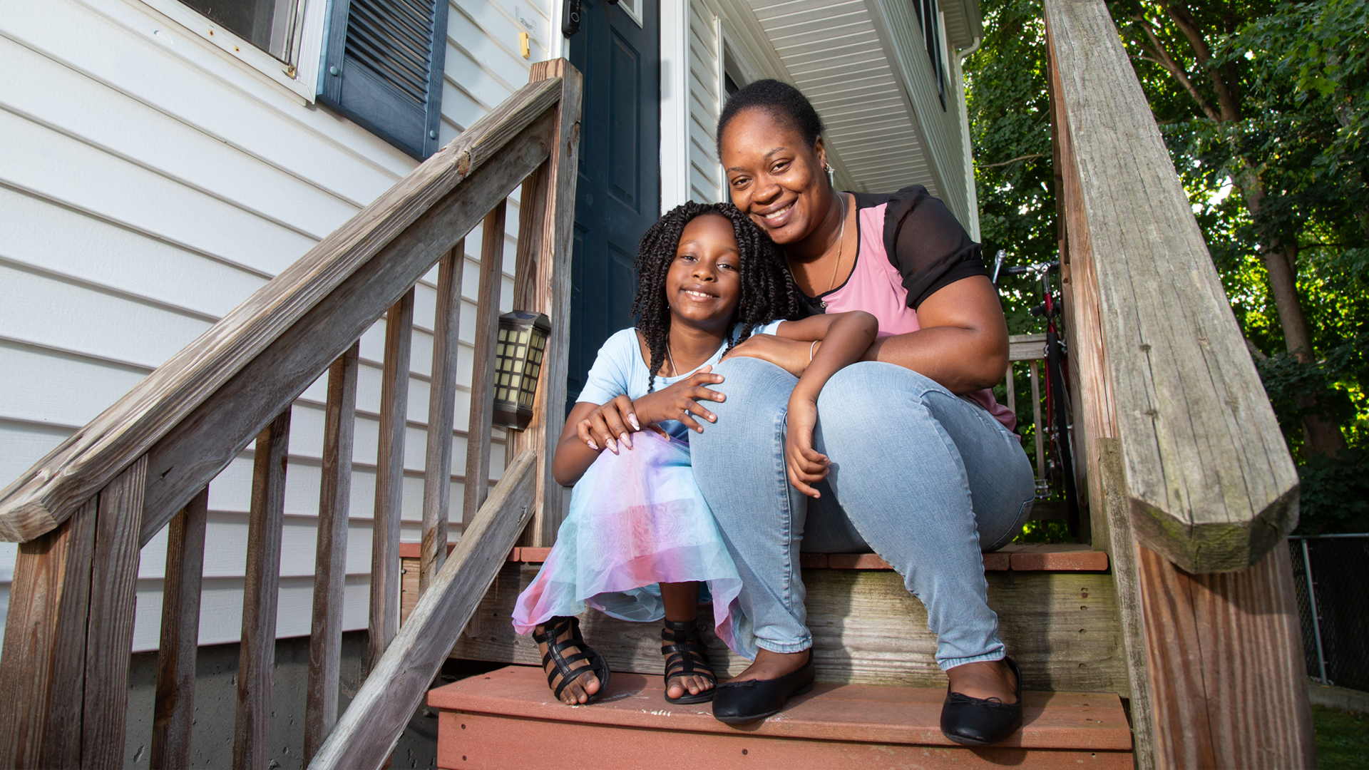 smiling mother and her young daughter sitting close to each other on the steps leading to the porch and front door of their home