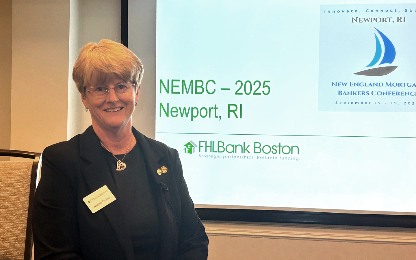 Picture featuing smiling woman standing on the side of conference sinage for the 2025 New England Mortage Bankers Conference