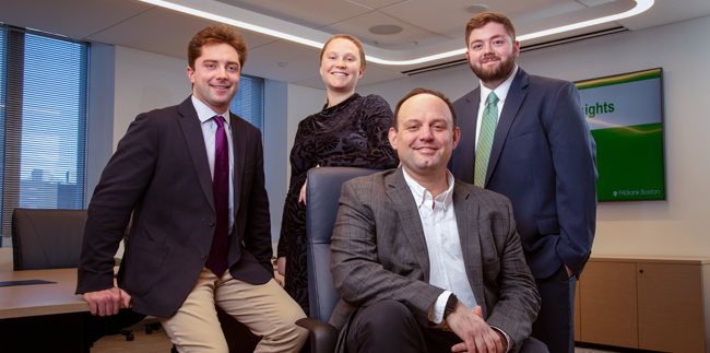 Four people in a room. One man is seated in a chair in the foreground. Behind him, are three others: a man leaning on a table to the left, a woman standing directly behind the seated man, and another man standing to the right.
