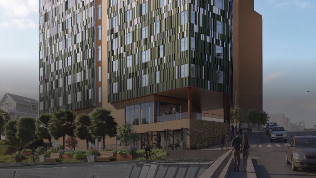 a multi-story apartment building facing a walkway, with a cyclist and pedestrians walking by and a busy road with cars driving past on one side