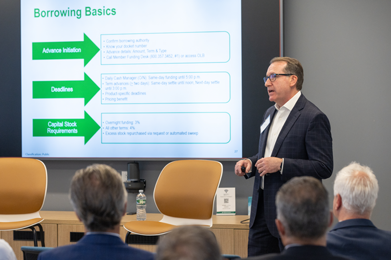 A presenter stands at the front of a conference room delivering a presentation to an audience seated in the foreground. A large screen displays a slide titled “Borrowing Basics,” featuring green arrows and sections labeled “Advance Initiation,” “Deadlines,” and “Capital Stock Requirements,” with supporting bullet points. The presenter is wearing a dark blazer and holding a presentation remote while speaking. Modern conference room furnishings, including tan chairs and a water bottle on a table, are visible beneath the screen.