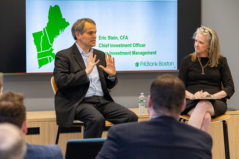 Two speakers seated in front of a presentation screen; the male speaker is gesturing while speaking, while the female speaker listens with a notebook on her lap. Audience members are visible in the foreground. The screen behind them displays a presentation slide with a green map graphic and text about investment management at FHLBank Boston.