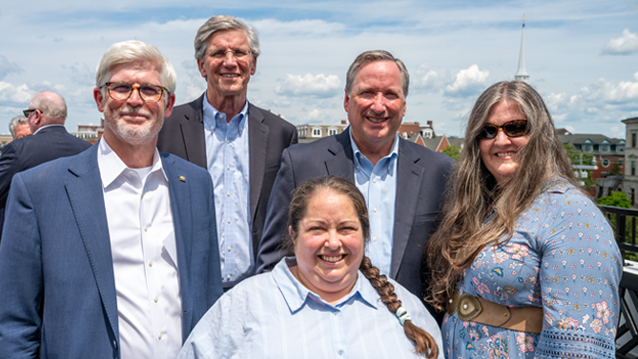 three men wearing suit jackets and two women standing next to each other outdoors while smiling with a of downtown Portsmouth in the background